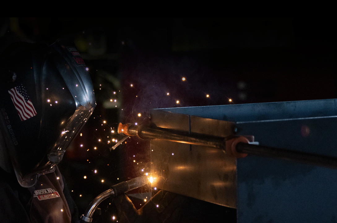 A welder wearing a helmet with an American flag is welding a part of a grill as orange sparks fly