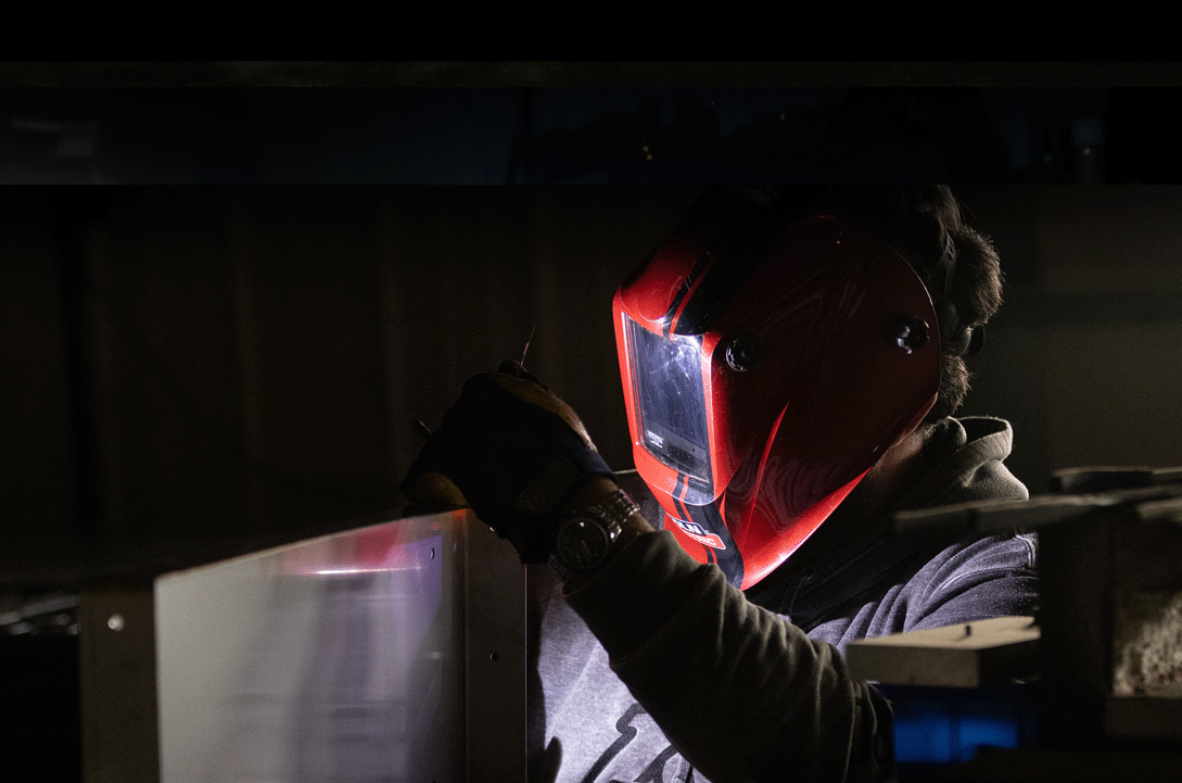 Chris Engelbrecht with red welding mask on is holding part of a grill while welding with blue light reflection
